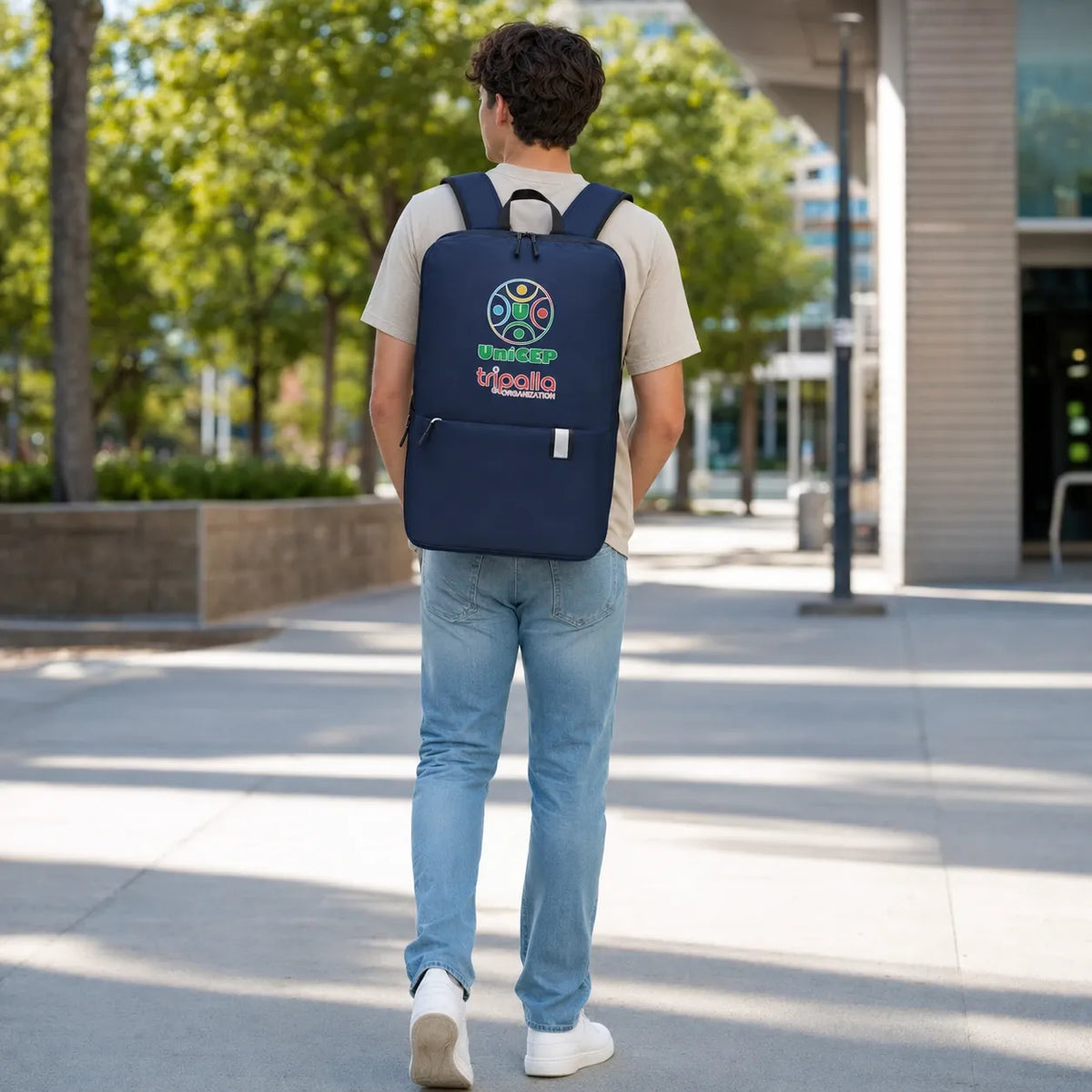 A man viewed from behind walking on a sunny outdoor campus walkway, wearing a navy blue backpack with a colorful circular logo and the text 'UNIVERSITY OF CALIFORNIA' on the back.