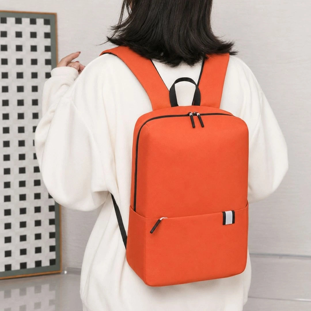 A person wearing a white sweatshirt is shown from behind carrying a bright orange backpack with black trim and straps in an indoor setting.