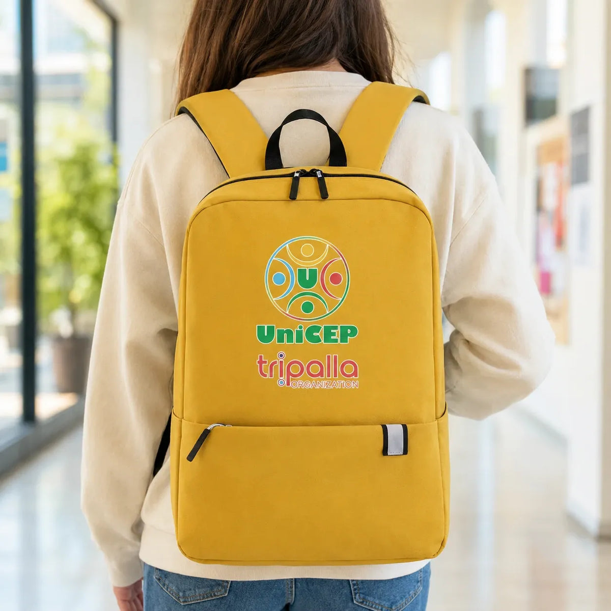 A person viewed from behind wearing a bright yellow backpack with a colorful 'uniCEP tripalla' logo on the back, standing in a brightly lit indoor hallway.