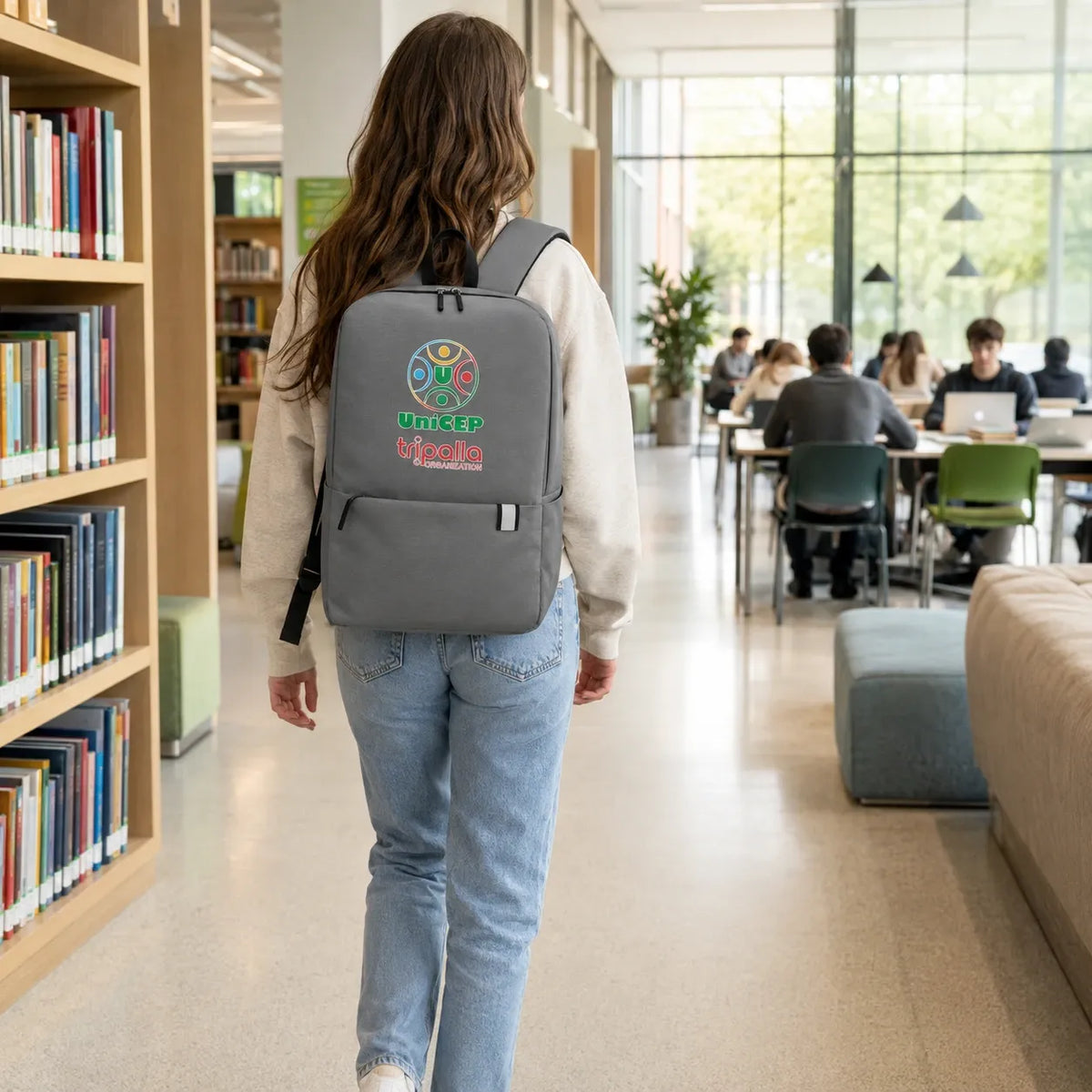 A woman walking away in a library or study lounge, wearing a gray backpack with a colorful circular graphic and the text 'UNICHD' on the back. Bookshelves are visible on the left, and other people are seated in the background near large windows.